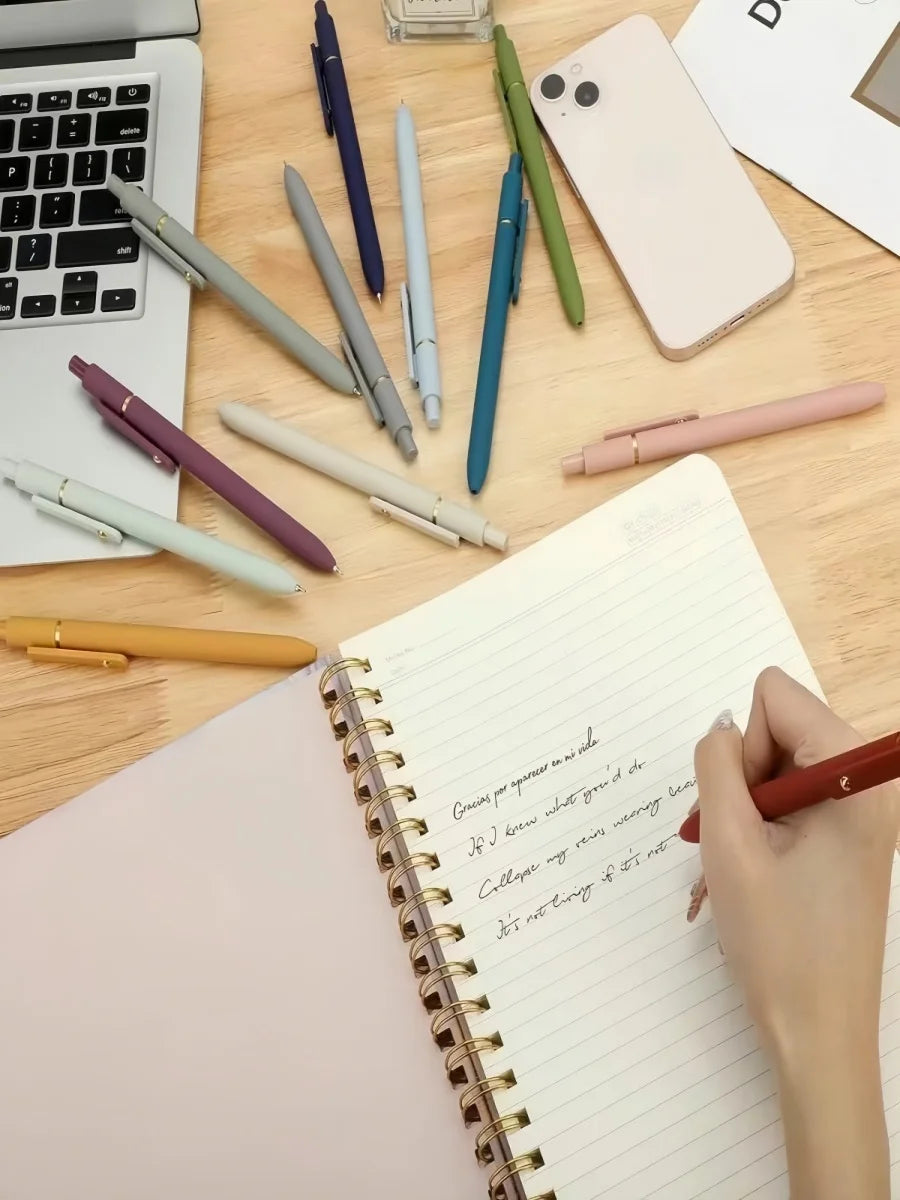 A collection of Morandi Gel Pens in various colors and shades laid out on a wooden desk next to a notebook.