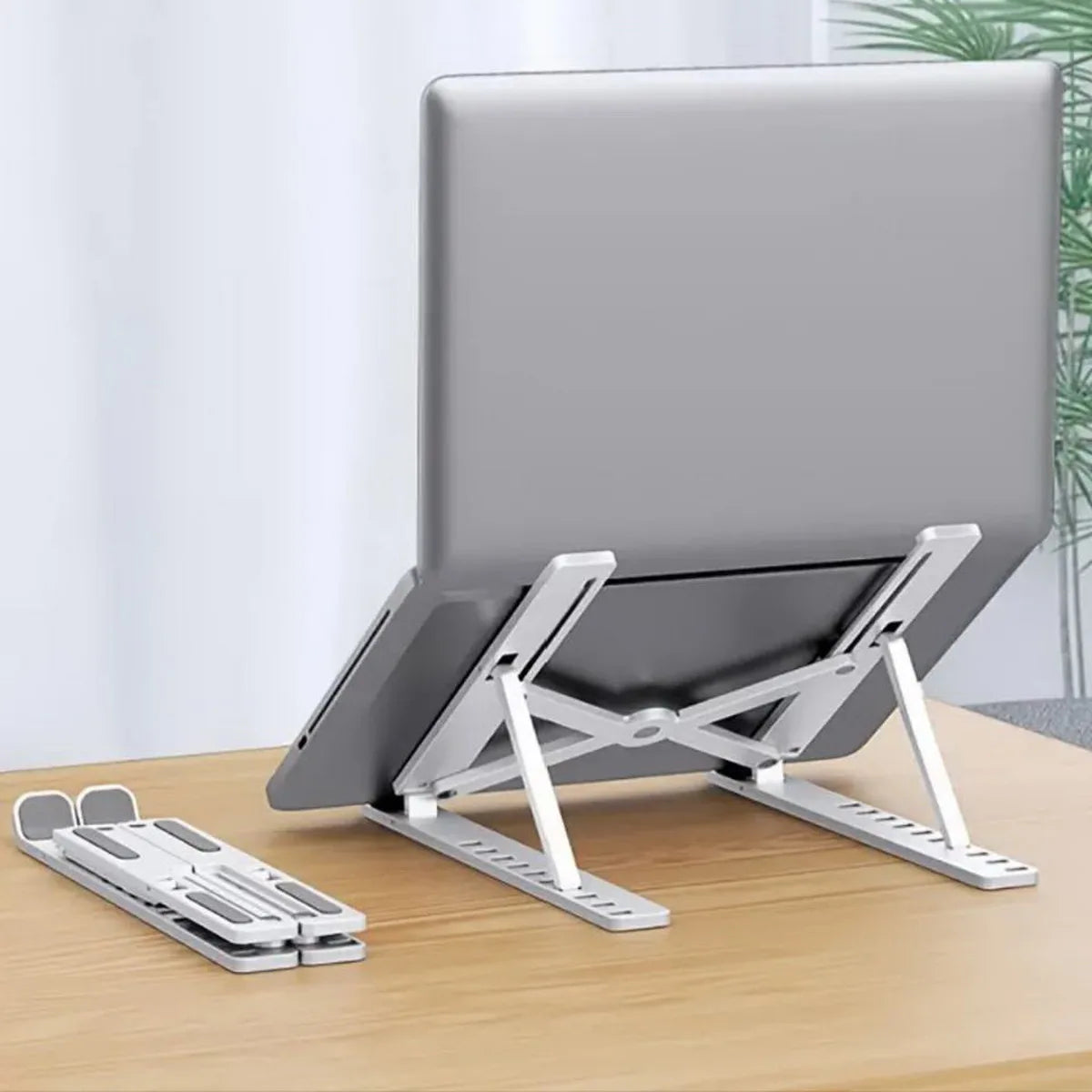 A silver portable folding laptop stand is shown on a wooden desk, ready to support a laptop.