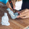 A person uses an electric foam cutting tool to carve or shape a light blue, sculpted piece of material on a wooden surface.