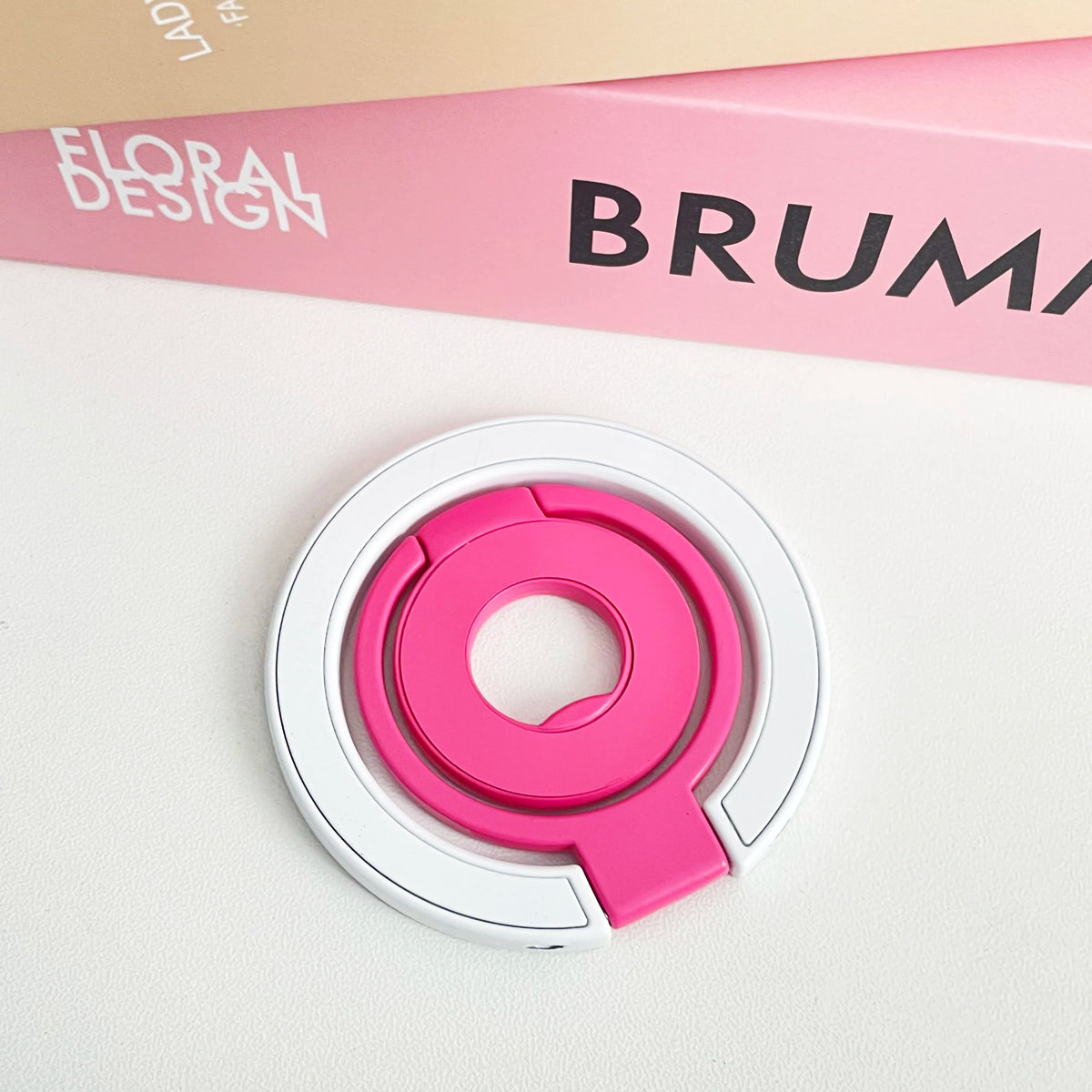 A pink magnetic phone grip and stand with a foldable ring is shown on a white surface.