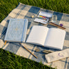 A white A5 dot grid notebook with a gold foil botanical cover lies open on a blue and white plaid cloth outdoors.