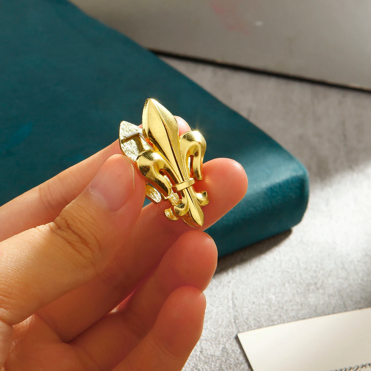 A hand holds a pair of decorative gold iris-shaped metal binder clips against a teal background.
