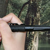 A hand holds a black tactical self-defense pen with a glass breaker tip against a tree trunk in a forest setting.