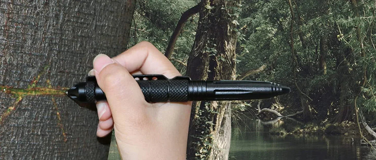 A hand holds a black tactical self-defense pen with a glass breaker tip against a tree trunk in a forest setting.