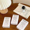 A white folding phone stand is shown on a wooden table next to a rolled-up paper and a light fixture.