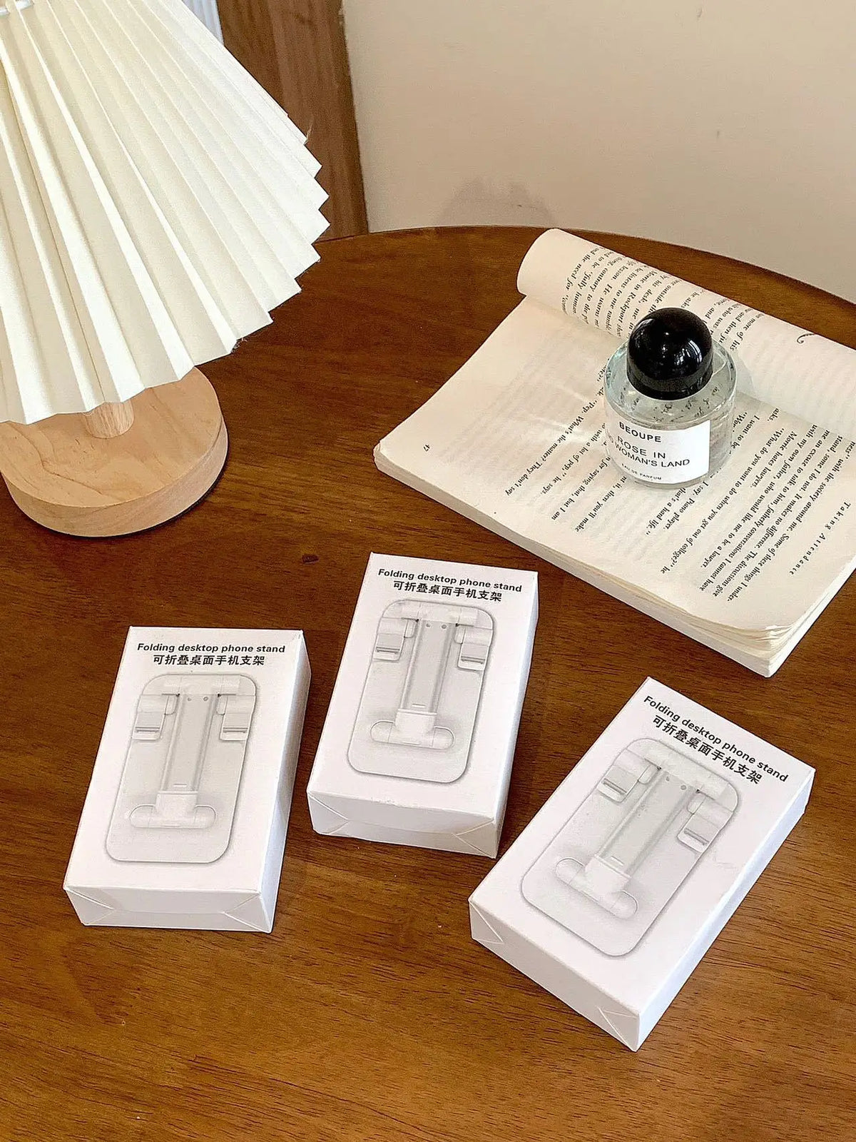 A white folding phone stand is shown on a wooden table next to a rolled-up paper and a light fixture.