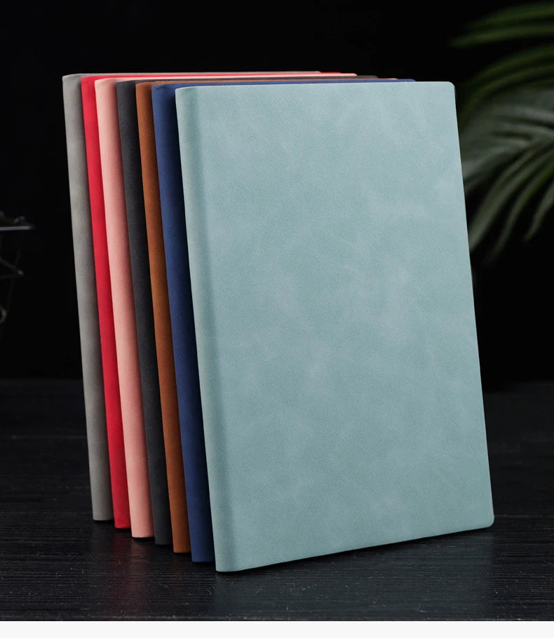 A stack of premium A5 leather journals in various muted colors, including red, pink, brown, and blue, displayed on a dark sur