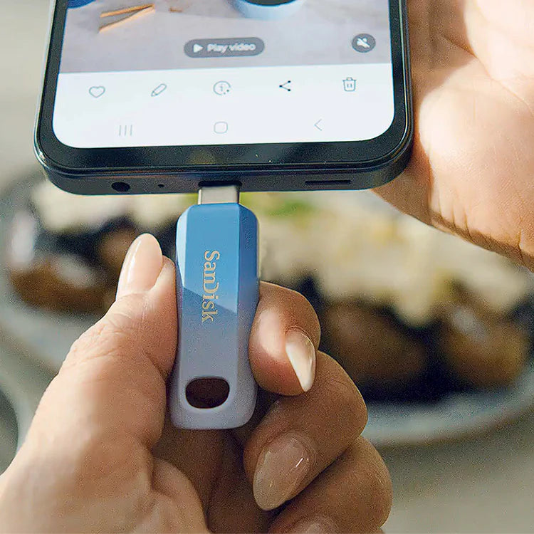 A hand holds a blue SanDisk flash drive, connecting it to a smartphone screen displaying a video player.