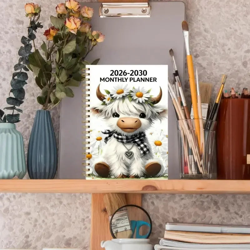 A monthly planner cover featuring a cute white bull with horns and a floral wreath, displayed on a wooden shelf.
