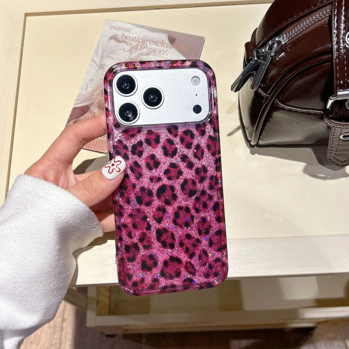 A close-up of a phone case with a leopard print design and pink sequins is held by a hand next to a brown leather bag.