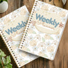 A spiral notebook weekly planner with floral patterns and a light beige cover is shown on a wooden surface.