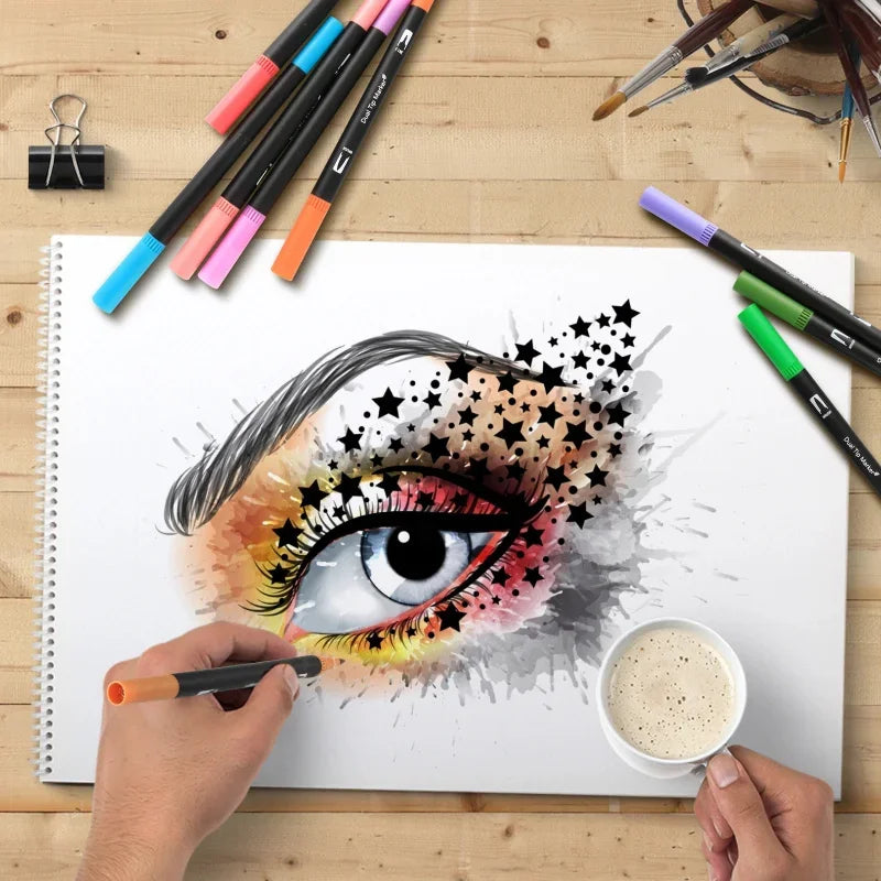 A hand is using a dual-tip brush marker from a set to draw an eye design on a white notepad.