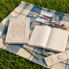 A white A5 dot grid notebook with a gold foil botanical cover lies on a blue and white plaid cloth outdoors.