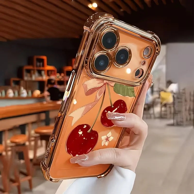 A hand holds an orange-toned iPhone case featuring a cherry and bow pattern with an electroplated finish.