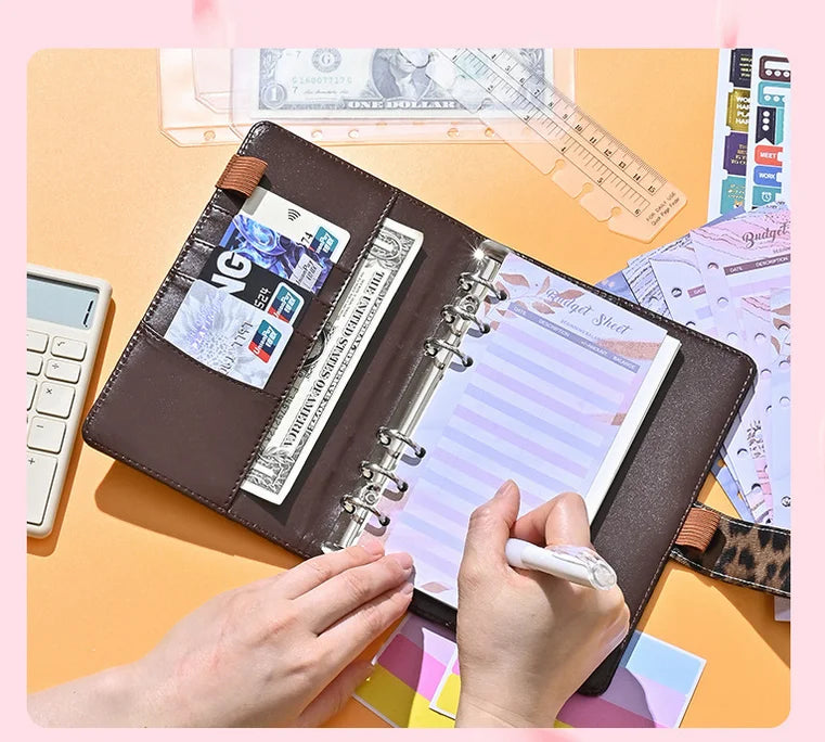 A hand is writing in a brown leopard print A6 binder with a clear plastic cover and loose leaf sheets on an orange surface.