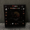 Schwarze Karte mit 'Happy Halloween' und Silhouette eines gruseligen Hauses vor orangem Mond.