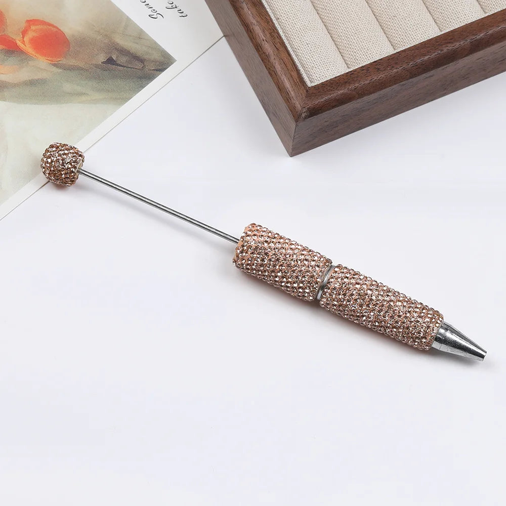 A long, silver pen accessory with a gold rhinestone ball and a textured beaded tip resting on a white surface.