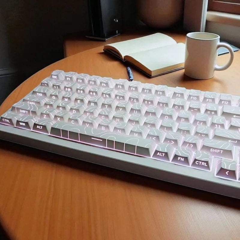 A close-up view of a light pink side-lit cherry profile mechanical keyboard resting on a wooden table next to a notebook and 