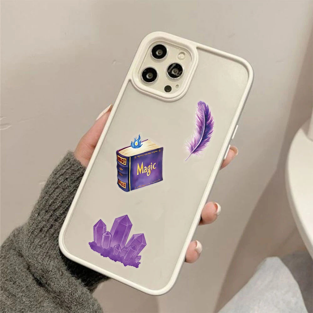 A hand holds a white smartphone decorated with purple magic witch stickers, including a book, an amethyst crystal, and a feat