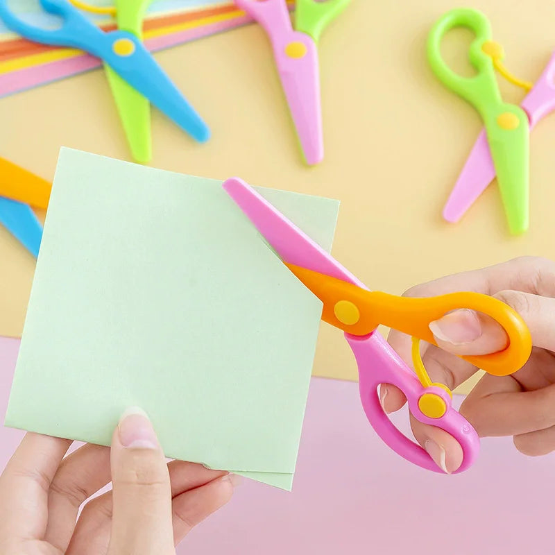 A hand is holding a pink and orange pair of plastic scissors, with several other colored craft scissors visible in the backgr