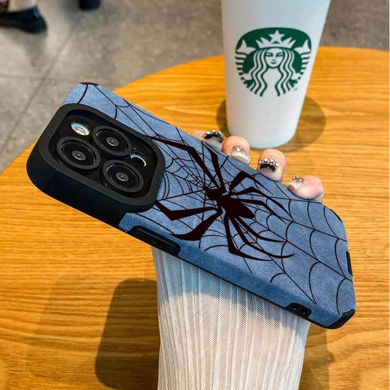 A dark blue phone case featuring a black spider web design, held by a person against a wooden table with a Starbucks cup in t