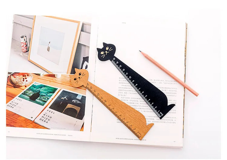 A wooden drawing ruler shaped like a cute cartoon cat is displayed on white paper with framed artwork.