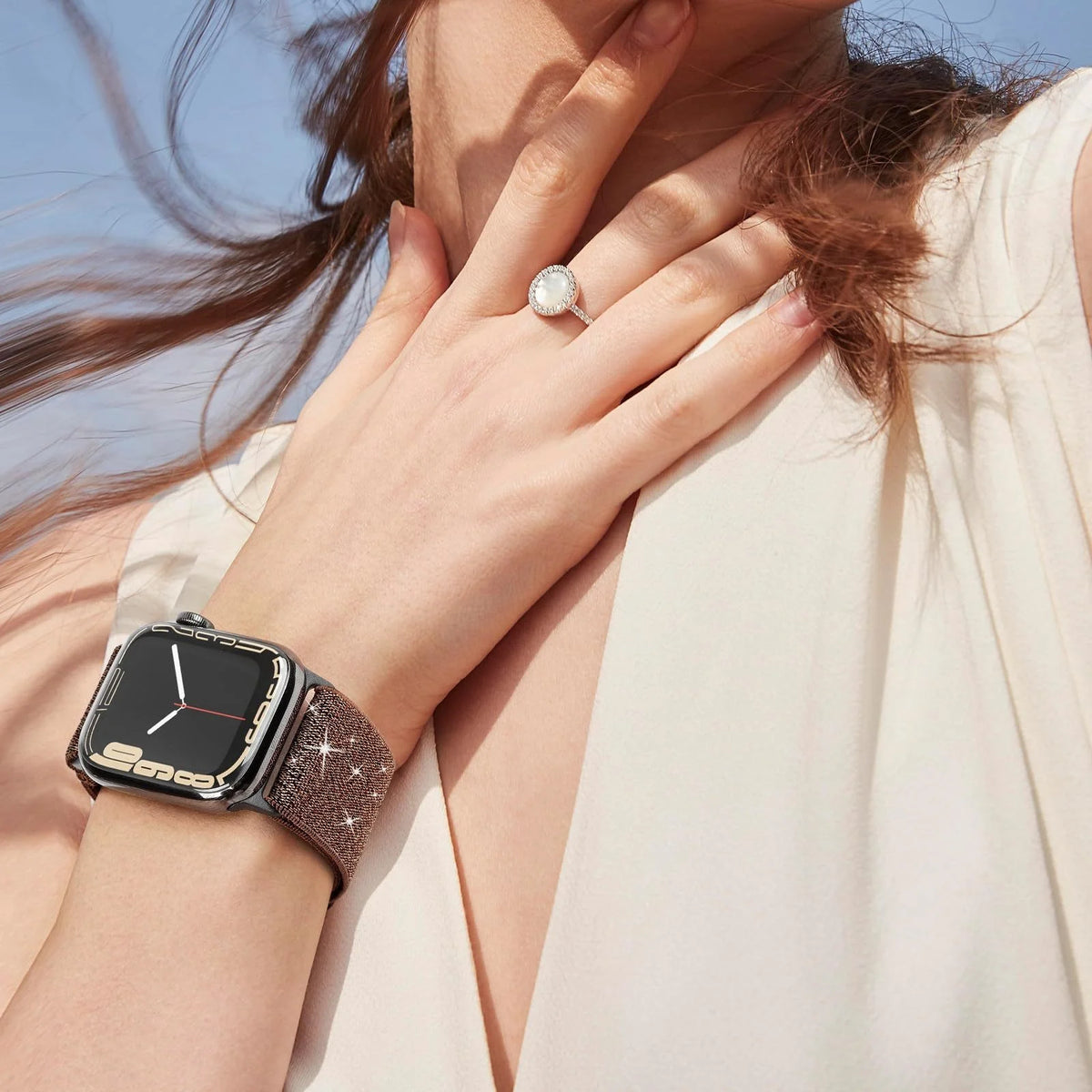 A close-up of a hand wearing a silver ring and a black Apple Watch with a glitter nylon strap against a light background.