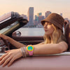 A woman wearing a brown hat and gold bracelets rests her arm on the door frame of a car, displaying a pink ceramic watch.