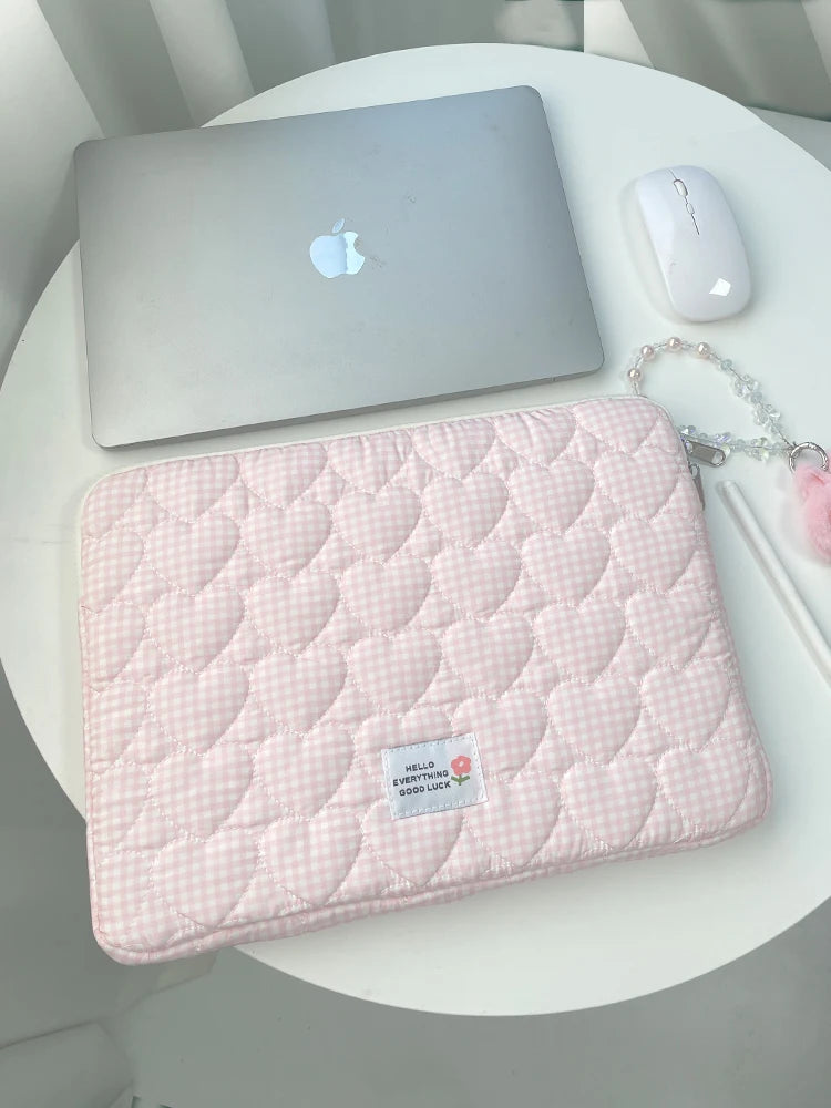 A pink quilted laptop sleeve with a white gingham pattern rests on a white table next to an Apple laptop and mouse.
