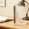 A close-up of a leopard print retractable ballpoint pen resting on a wooden table next to a desk lamp and colored sticky note