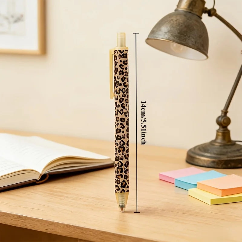 A close-up of a leopard print retractable ballpoint pen resting on a wooden table next to a desk lamp and colored sticky note