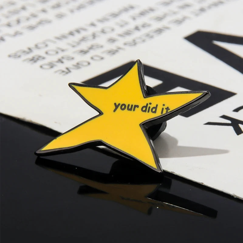 A yellow star enamel pin with the text your did it is placed on a dark surface.