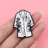 A white enamel pin shaped like a doctor's coat featuring floral embroidery and a pencil detail, held by a hand.