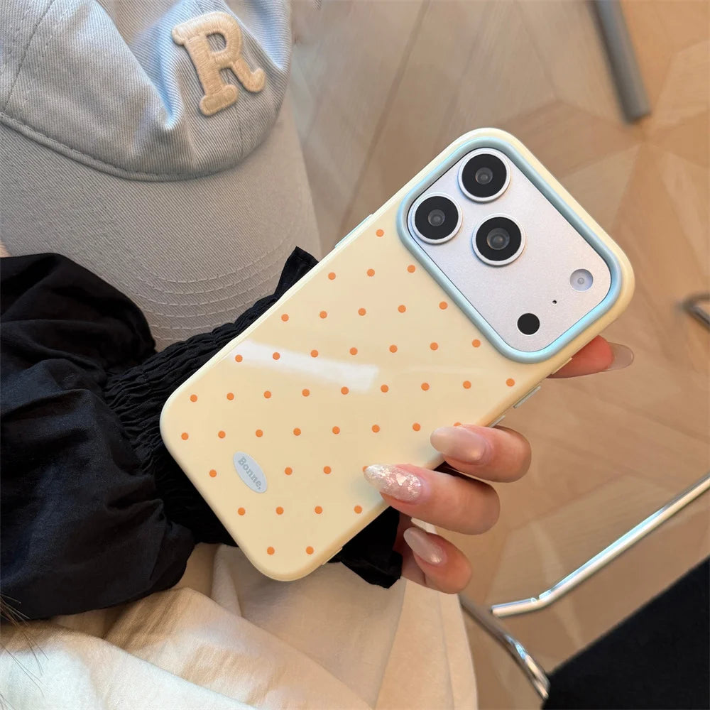A hand holds a light yellow phone case with white polka dots and a clear ring, next to a gray hat.