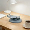 A gray foldable desktop phone stand resting on a wooden table next to a white lamp.