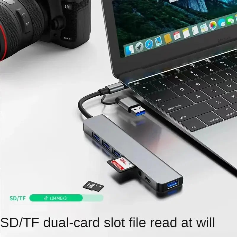 A silver USB hub docking station with two SD/TF cards is shown next to a laptop and a camera lens.