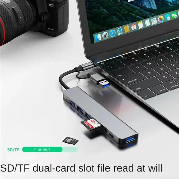 A silver USB hub docking station with two SD/TF cards is shown next to a laptop and a camera lens.