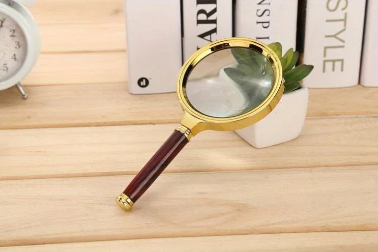 A gold-framed magnifying glass with a dark handle rests on a wooden surface next to a stack of books.