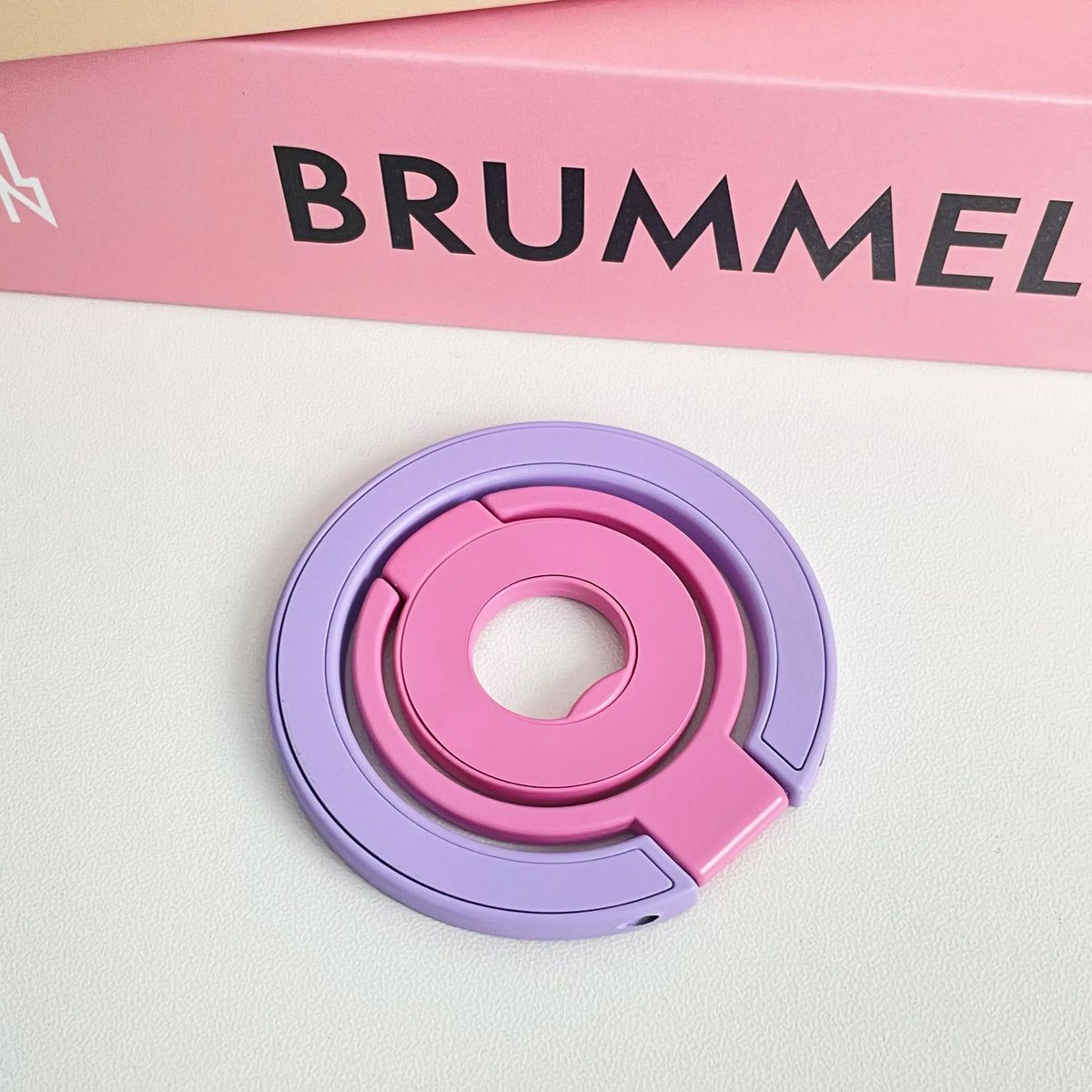 A pink and purple magnetic phone grip and stand with a foldable ring resting on a white surface.