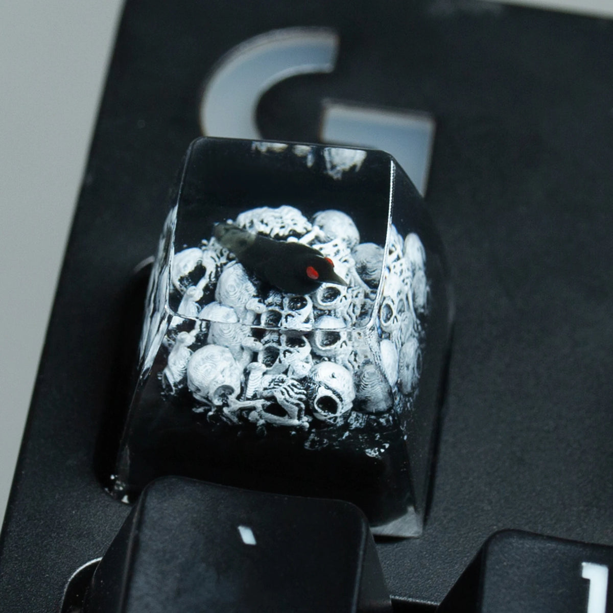 A close-up view of a black resin keycap featuring a detailed design of skulls and a raven's eye.