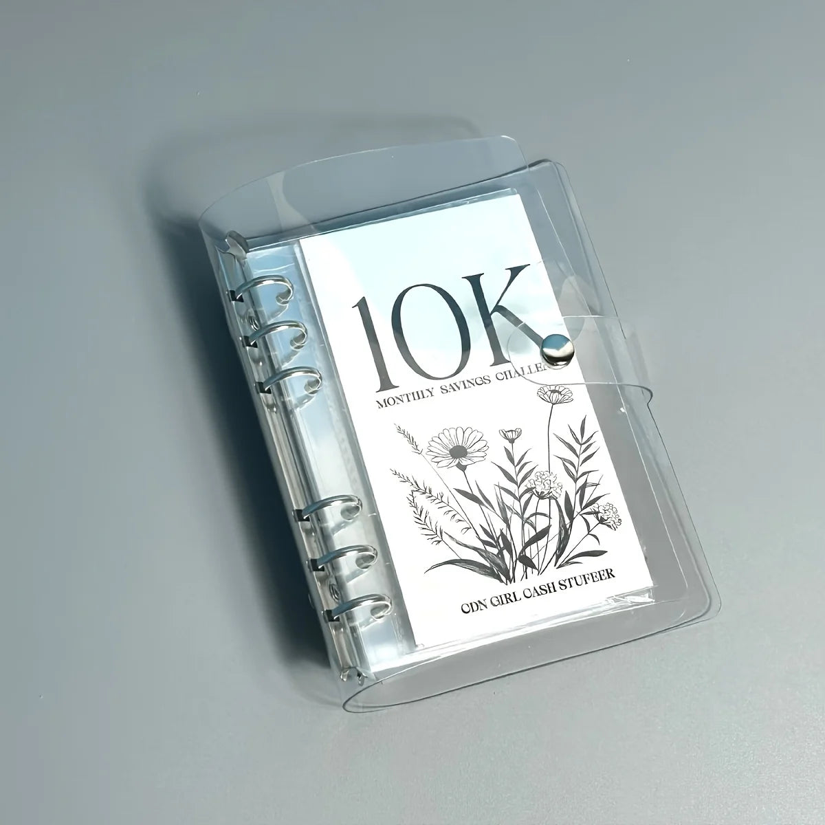 A clear plastic binder showing a monthly savings challenge with floral illustrations and the name Cin Girl Cashi Stufeer