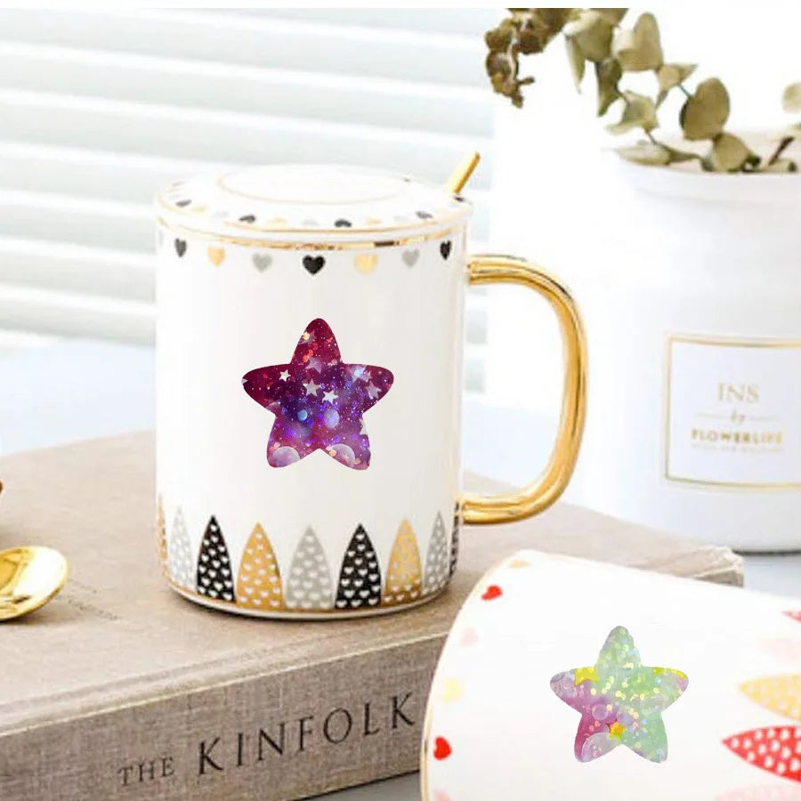 Weiße Keramiktasse mit goldfarbener Henkel, verziert mit lila Glitzersternen und goldenen Akzenten.