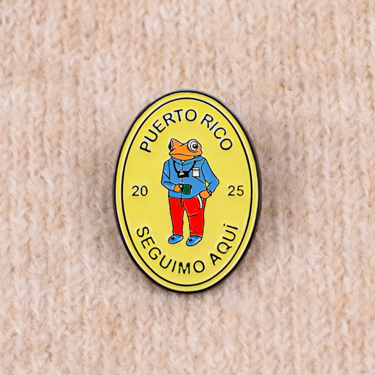 A yellow enamel pin featuring an orange frog wearing a blue shirt and red pants, with the text Puerto Rico and Seguimo Aquí.