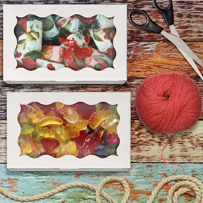 Two white boxes display colorful candies and cookies next to a ball of red yarn and scissors on a wooden surface.