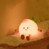 A glowing peach-shaped night light with a smiling face and green stem resting on a soft fabric.