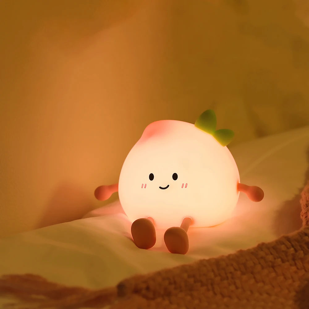 A glowing peach-shaped night light with a smiling face and green stem resting on a soft fabric.
