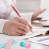 A hand is writing with a pink mechanical pencil on paper next to a set of pastel colored pencils.