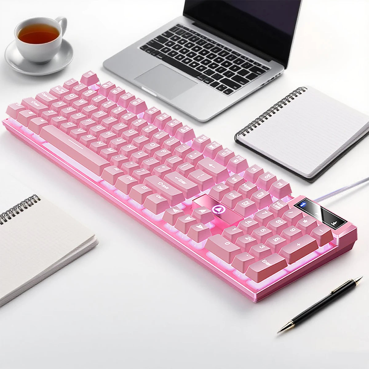 A pink low profile wired gaming keyboard with a numpad is displayed next to a laptop and a notebook.