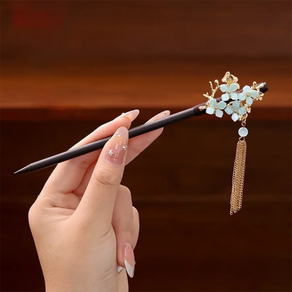Une main tient un pinceau noir avec des accessoires pour cheveux floraux dorés, des fleurs bleu clair et une chaîne dorée pen