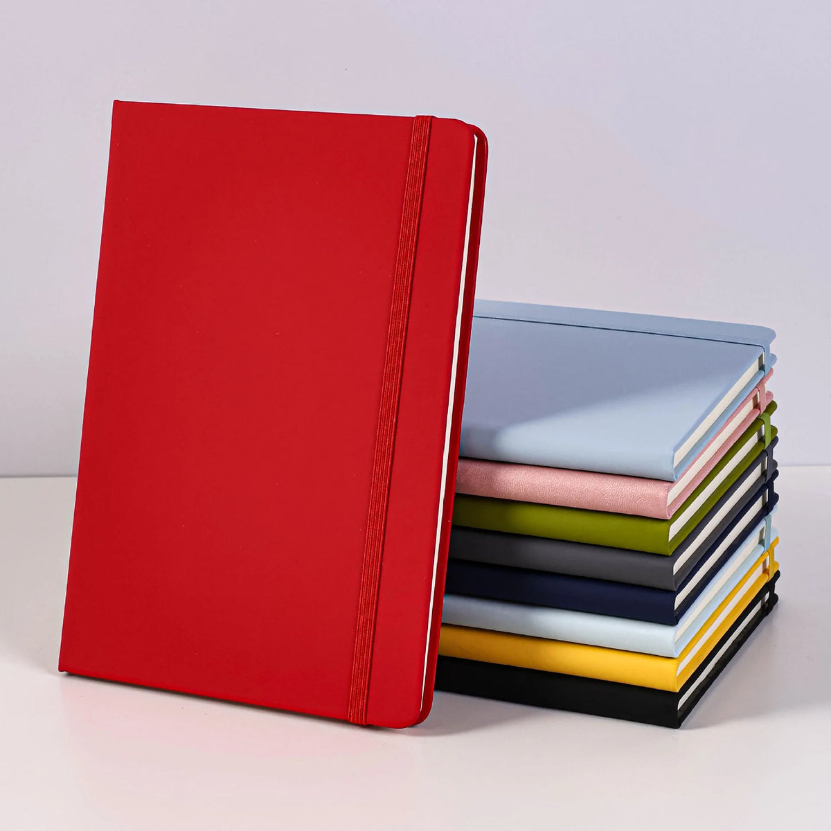 A red A5 hardcover lined notebook next to a stack of colored notebooks.